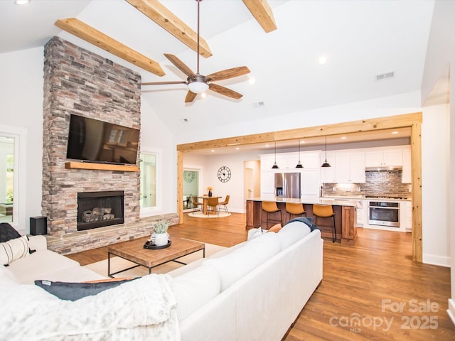 living room with a stone fireplace, ceiling fan, light hardwood / wood-style flooring, and vaulted ceiling with beams