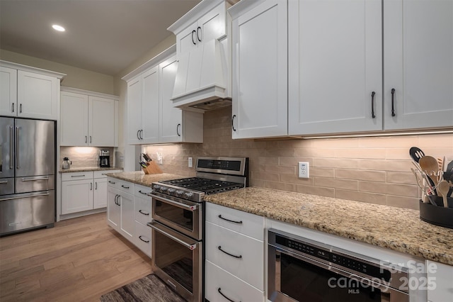 kitchen featuring light stone countertops, backsplash, high quality appliances, white cabinets, and light wood-type flooring