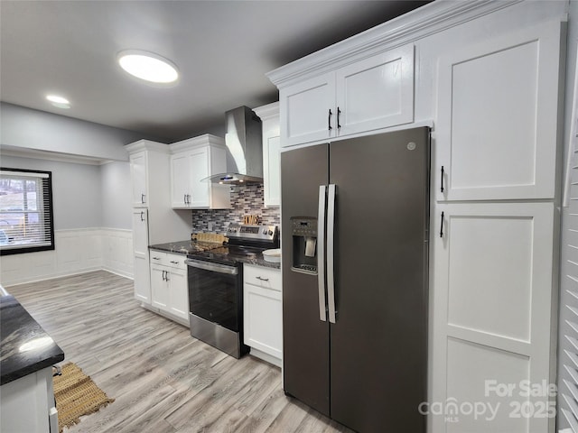 kitchen featuring white cabinets, appliances with stainless steel finishes, wall chimney exhaust hood, dark stone counters, and tasteful backsplash