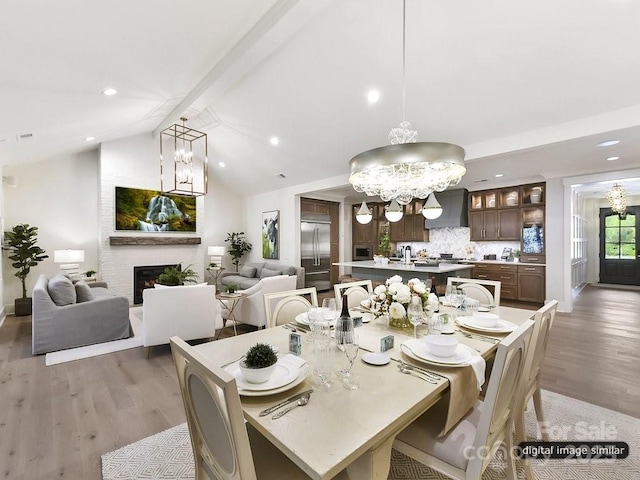 dining space with vaulted ceiling with beams, a large fireplace, light wood-type flooring, and an inviting chandelier