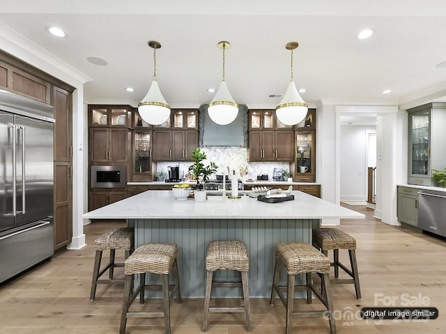 kitchen with built in appliances, custom exhaust hood, a center island with sink, and decorative light fixtures