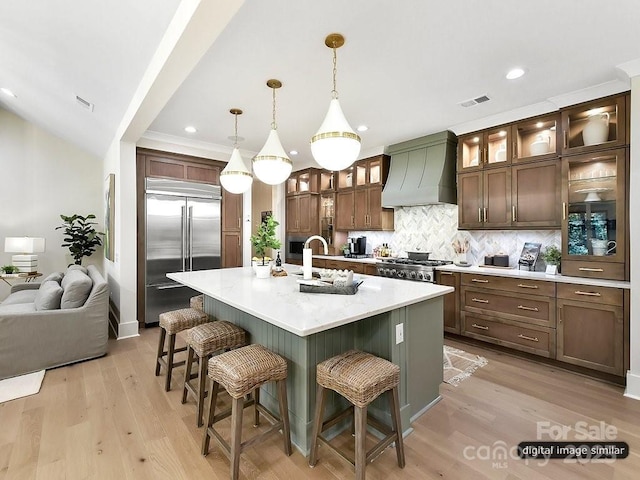 kitchen with sink, stainless steel built in refrigerator, premium range hood, decorative light fixtures, and a kitchen island with sink