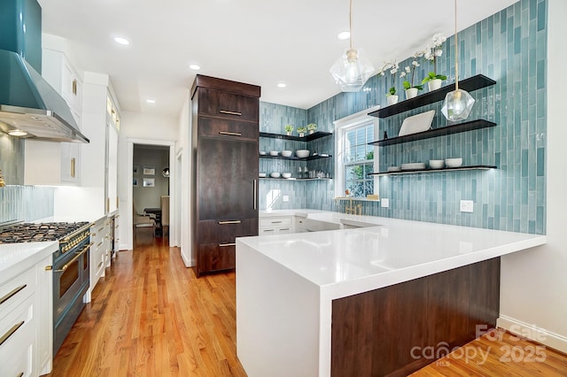 kitchen with dark brown cabinetry, wall chimney exhaust hood, hanging light fixtures, range with two ovens, and white cabinets