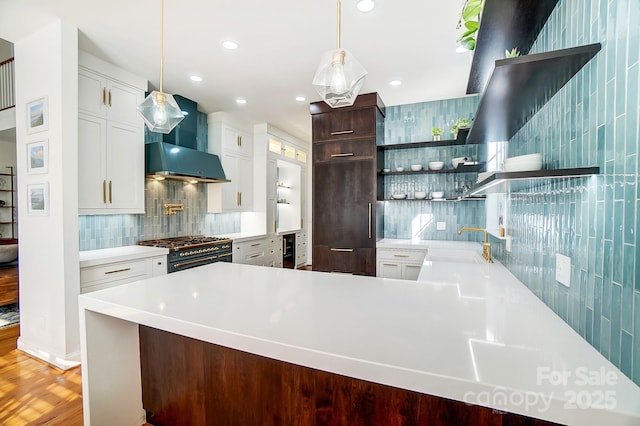 kitchen featuring high end stainless steel range oven, sink, wall chimney range hood, white cabinets, and hanging light fixtures