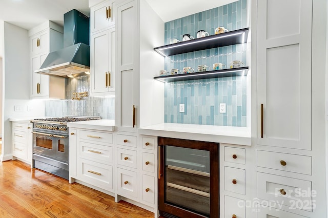 kitchen with light wood-type flooring, wall chimney range hood, range with two ovens, white cabinets, and wine cooler