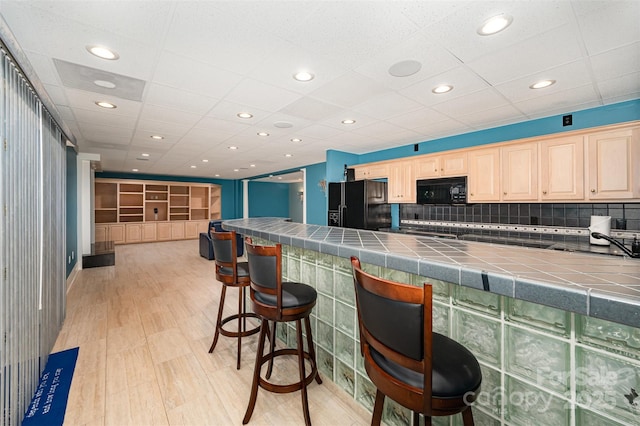 kitchen featuring tile counters, backsplash, a drop ceiling, a breakfast bar, and black appliances