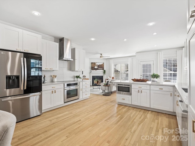 kitchen featuring wall chimney exhaust hood, light hardwood / wood-style flooring, stainless steel appliances, decorative backsplash, and white cabinets