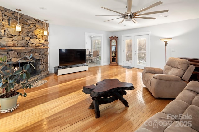 living room with ceiling fan, a stone fireplace, light wood-type flooring, and french doors