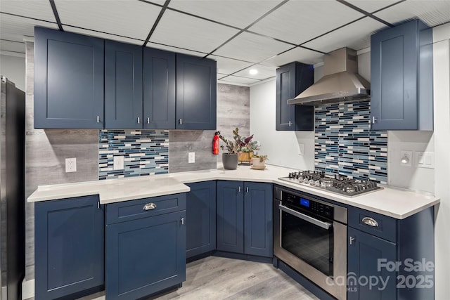 kitchen featuring wall chimney exhaust hood, blue cabinets, light hardwood / wood-style flooring, appliances with stainless steel finishes, and backsplash