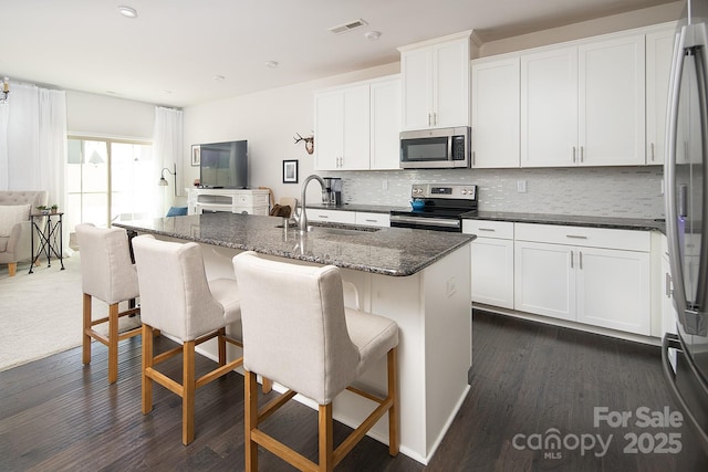 kitchen featuring stainless steel appliances, a kitchen island with sink, sink, dark stone countertops, and white cabinets