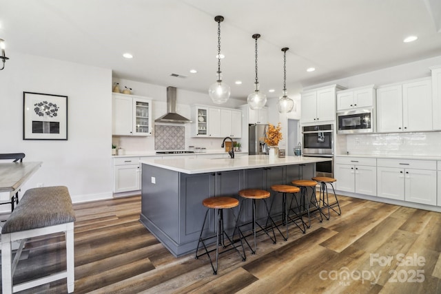 kitchen featuring stainless steel appliances, a center island with sink, white cabinetry, and wall chimney exhaust hood