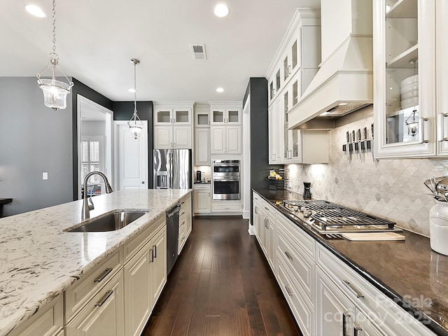 kitchen with appliances with stainless steel finishes, premium range hood, dark stone counters, pendant lighting, and sink