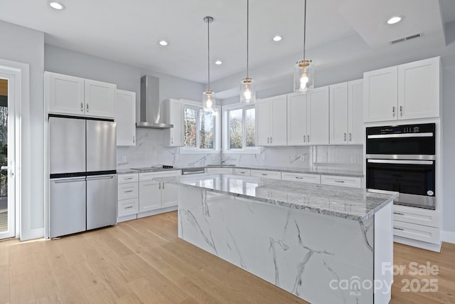 kitchen with a center island, wall chimney range hood, hanging light fixtures, appliances with stainless steel finishes, and white cabinetry