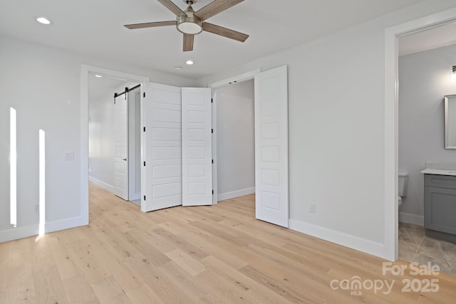 unfurnished bedroom featuring light wood-type flooring, a barn door, ensuite bath, and ceiling fan