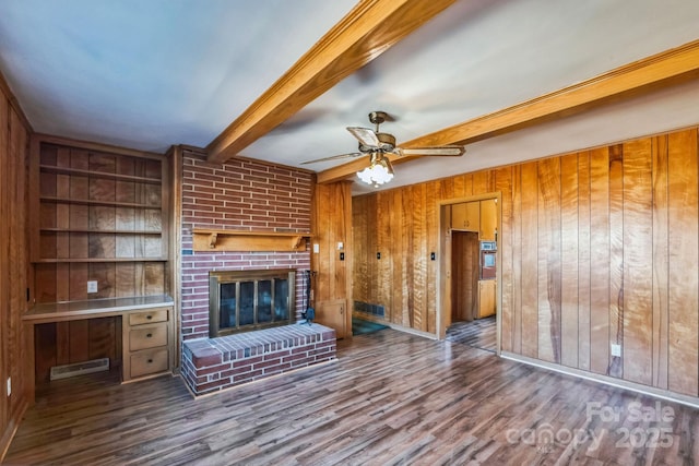 unfurnished living room with built in features, a fireplace, wooden walls, beamed ceiling, and ceiling fan