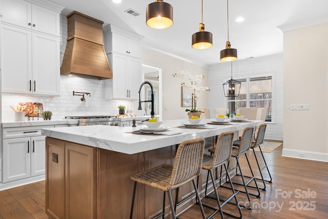 kitchen with white cabinets, hanging light fixtures, decorative backsplash, light stone countertops, and an island with sink