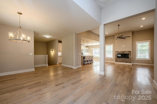 unfurnished living room with a stone fireplace, a healthy amount of sunlight, ceiling fan with notable chandelier, and light wood-type flooring