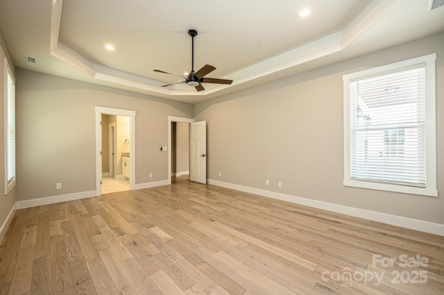 unfurnished bedroom featuring a raised ceiling, ensuite bath, ceiling fan, multiple windows, and light hardwood / wood-style floors