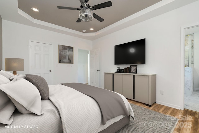 bedroom featuring ceiling fan, ensuite bathroom, a raised ceiling, and light wood-type flooring