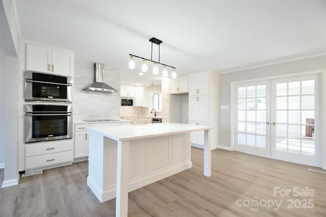 kitchen featuring double oven, white cabinetry, hanging light fixtures, and wall chimney range hood