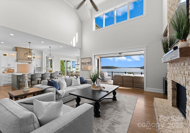 living room with a water view, a brick fireplace, ceiling fan, light wood-type flooring, and a towering ceiling