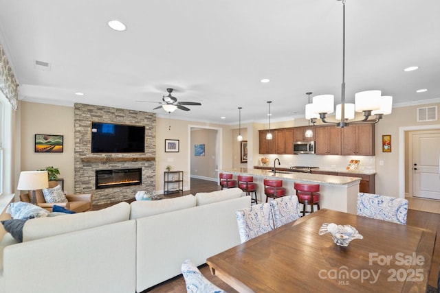 dining room featuring ceiling fan with notable chandelier, sink, a stone fireplace, and ornamental molding