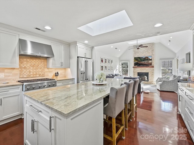 kitchen with vaulted ceiling with skylight, white cabinets, wall chimney exhaust hood, stainless steel appliances, and a stone fireplace