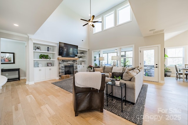 living room with a stone fireplace, ceiling fan, light wood-type flooring, a towering ceiling, and built in shelves