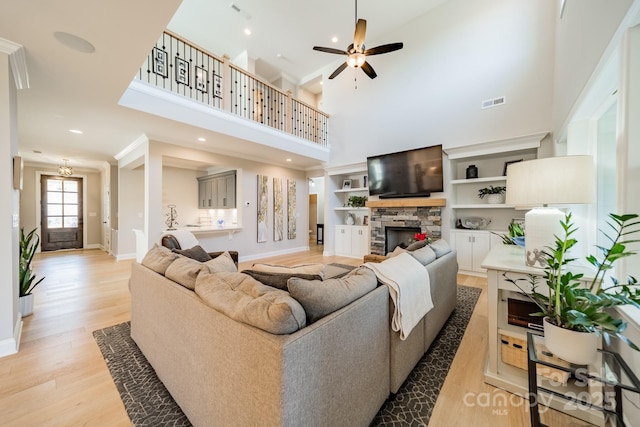 living room featuring ceiling fan, a towering ceiling, light wood-type flooring, built in features, and a stone fireplace