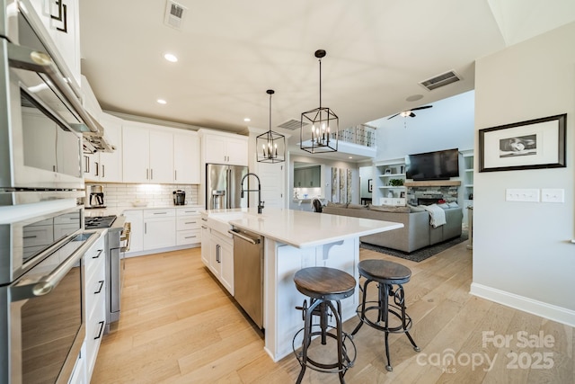 kitchen with ceiling fan with notable chandelier, white cabinets, stainless steel appliances, an island with sink, and hanging light fixtures