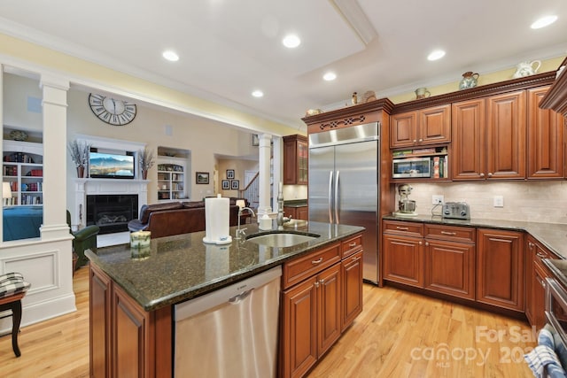 kitchen featuring sink, built in appliances, an island with sink, dark stone counters, and a fireplace