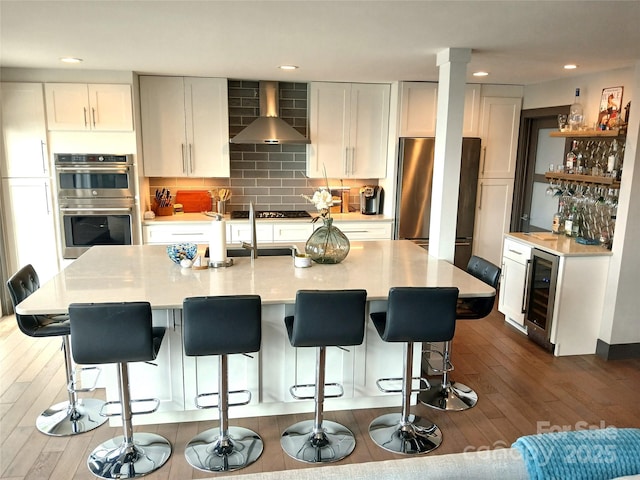 kitchen with wall chimney exhaust hood, white cabinetry, an island with sink, stainless steel appliances, and beverage cooler