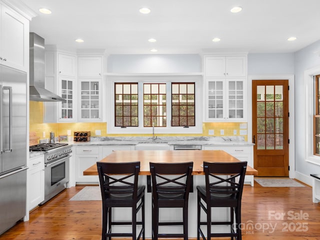 kitchen with high quality appliances, wall chimney range hood, a breakfast bar, light wood-type flooring, and white cabinets