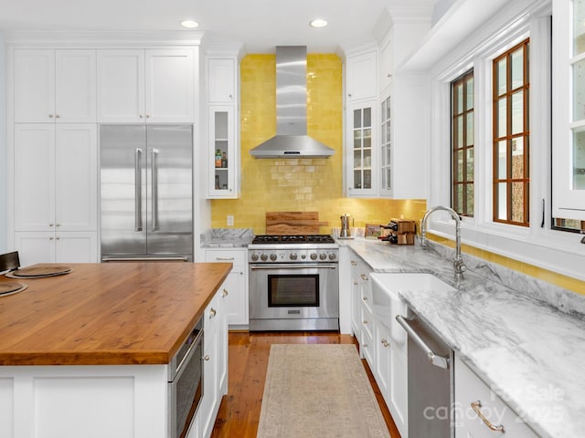 kitchen featuring built in appliances, light stone counters, wall chimney exhaust hood, white cabinetry, and tasteful backsplash