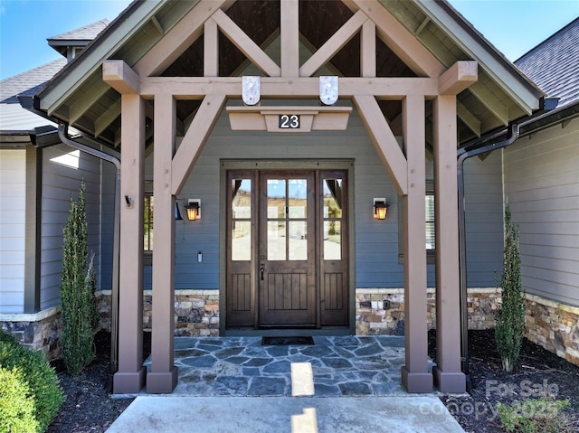 entrance to property featuring stone siding, roof with shingles, and covered porch