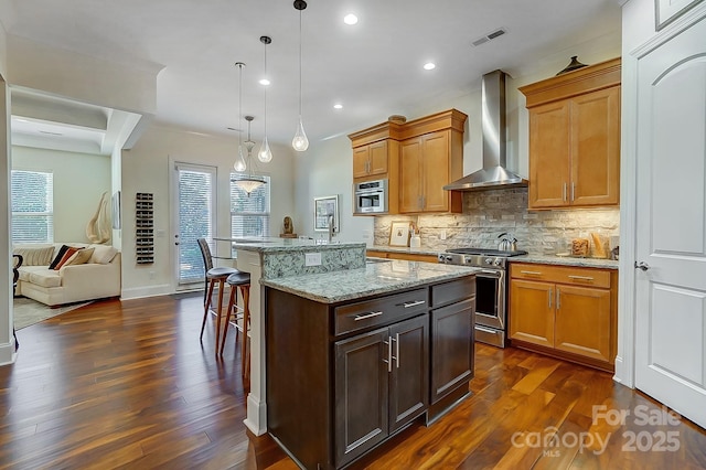 kitchen with hanging light fixtures, appliances with stainless steel finishes, a kitchen island, light stone countertops, and wall chimney range hood