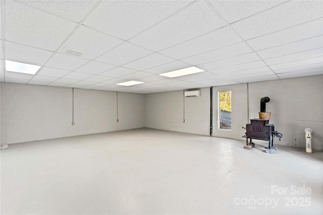 basement with a wall mounted air conditioner, a paneled ceiling, and a wood stove