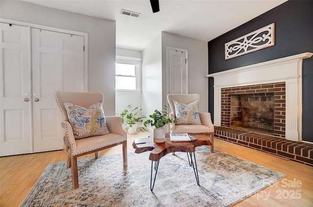 sitting room with a brick fireplace, wood finished floors, visible vents, and ceiling fan