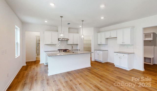 kitchen featuring light stone countertops, decorative light fixtures, a center island with sink, white cabinets, and light wood-type flooring