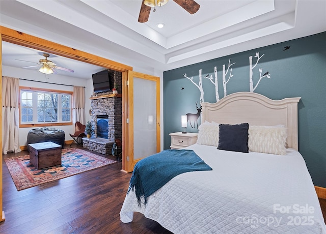 bedroom with dark hardwood / wood-style floors, a large fireplace, ceiling fan, and a tray ceiling