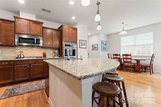 kitchen featuring a center island with sink, appliances with stainless steel finishes, hanging light fixtures, decorative backsplash, and light hardwood / wood-style flooring