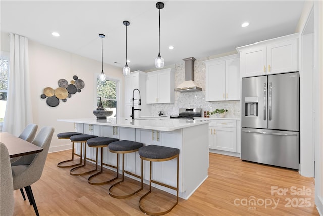 kitchen with a center island with sink, light wood finished floors, a sink, appliances with stainless steel finishes, and wall chimney exhaust hood