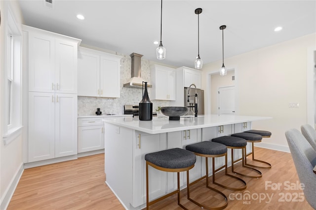 kitchen with a breakfast bar area, a kitchen island with sink, stainless steel appliances, white cabinetry, and wall chimney range hood