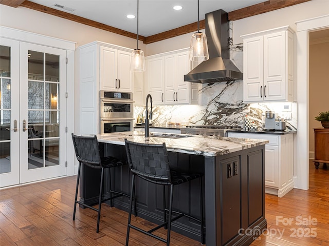 kitchen featuring dark stone countertops, a center island with sink, appliances with stainless steel finishes, white cabinets, and wall chimney range hood