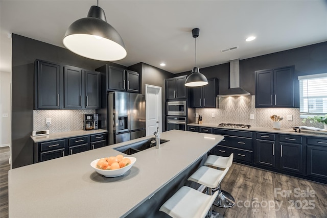 kitchen with decorative light fixtures, stainless steel appliances, a center island with sink, and wall chimney range hood