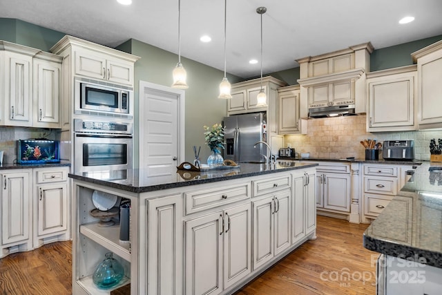 kitchen with a kitchen island with sink, stainless steel appliances, light wood-type flooring, hanging light fixtures, and tasteful backsplash