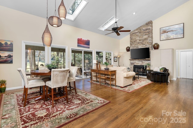 dining area with hardwood / wood-style floors, a skylight, a stone fireplace, high vaulted ceiling, and ceiling fan