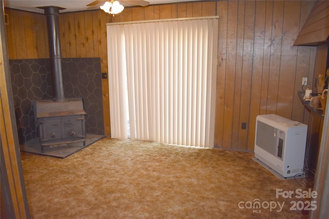 unfurnished living room featuring a wood stove, ceiling fan, carpet, heating unit, and wood walls