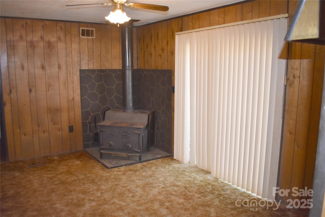 unfurnished living room with ceiling fan, a wood stove, wooden walls, and carpet flooring