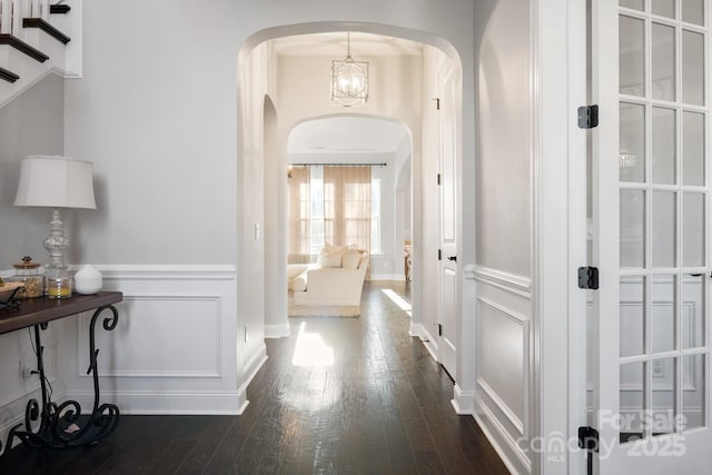 hallway featuring dark wood-type flooring, arched walkways, wainscoting, and a decorative wall